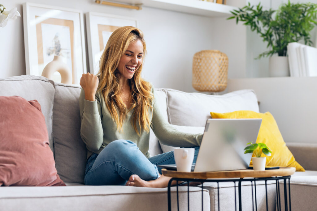 woman throwing her fist up in celebration while sitting on her couch and look at her laptop screen